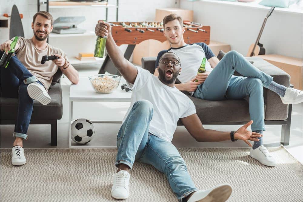 young men in living room watching football match and celebrating with beer bottles